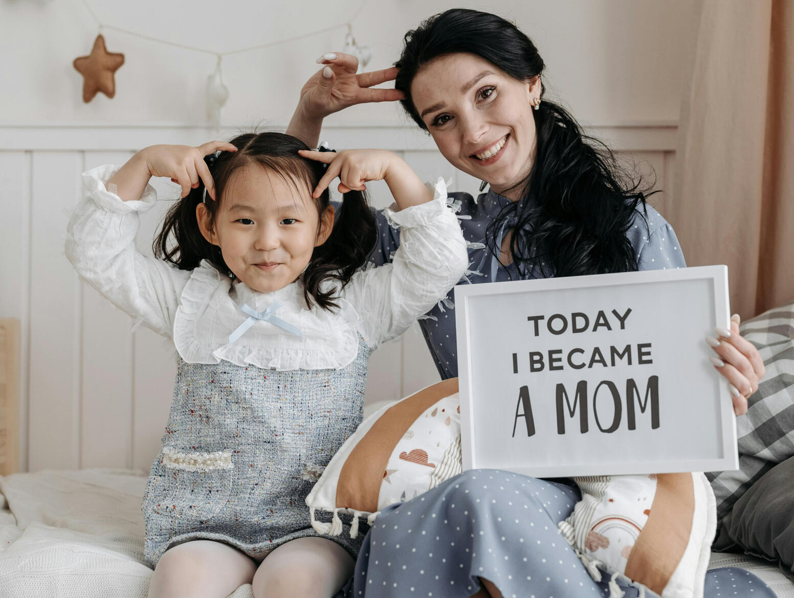 Mom and older daughter are sitting on a bed making silly faces holding a sign saying today I became A Mom