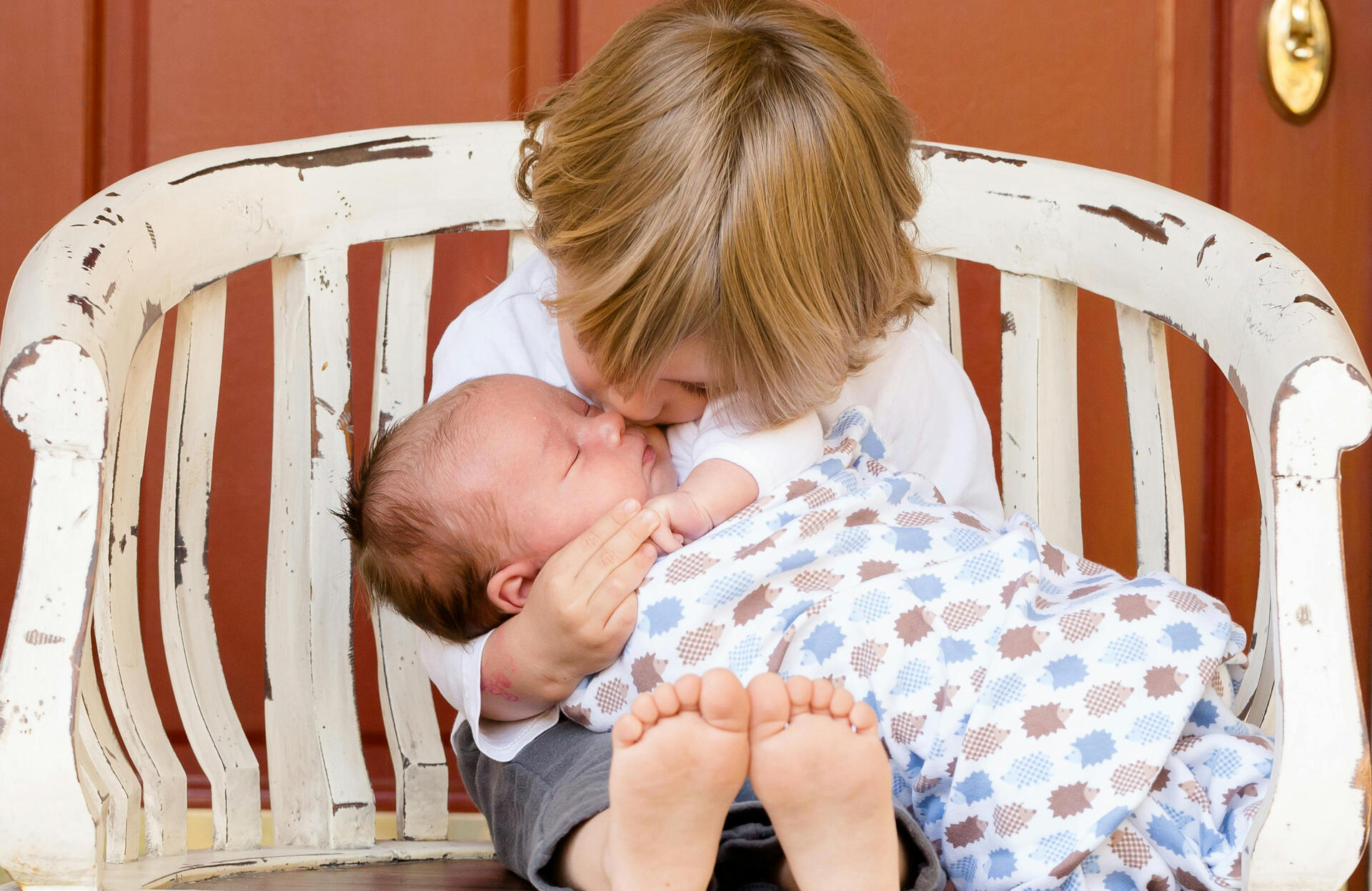 older sibling sitting in a chair holding a baby and kissing him on the forehead